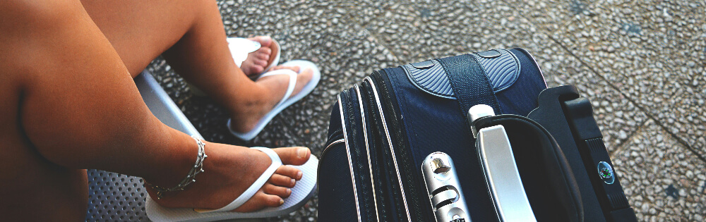 Reiseversicherungen Person sitzt auf einer Bank mit einem schwarzen Reisekoffer daneben und trägt weiße Flip-Flops sowie ein Fußkettchen.